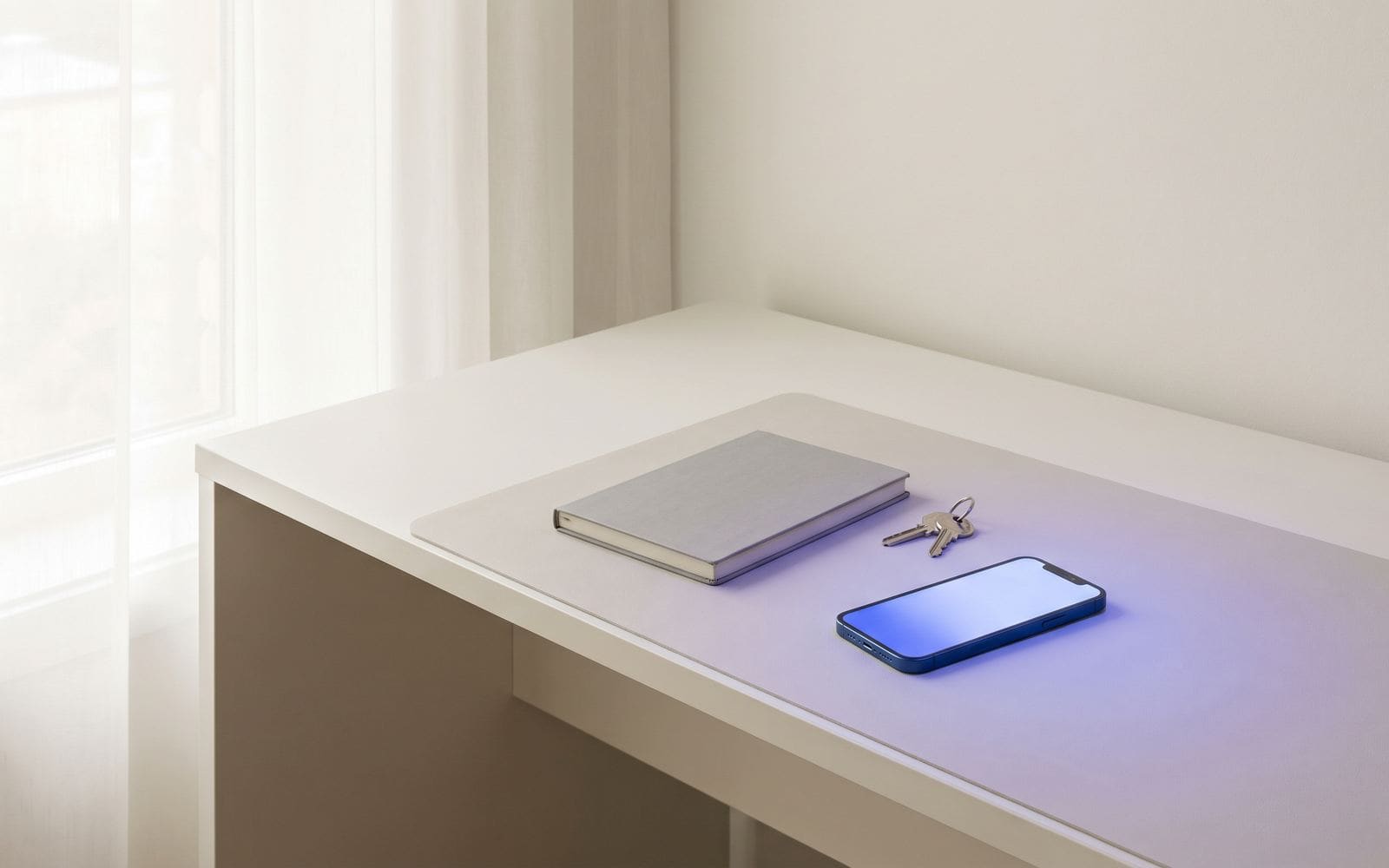 A tidy desk by a window with a smartphone, a notebook, and a set of house keys