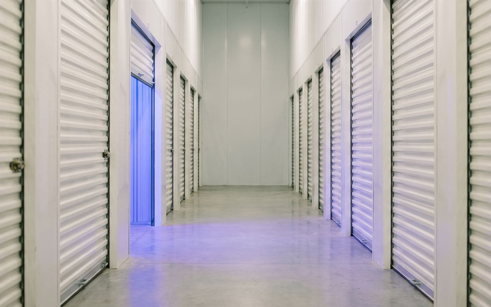 A corridor of roll-up storage unit doors, one partly open with blue light spilling out